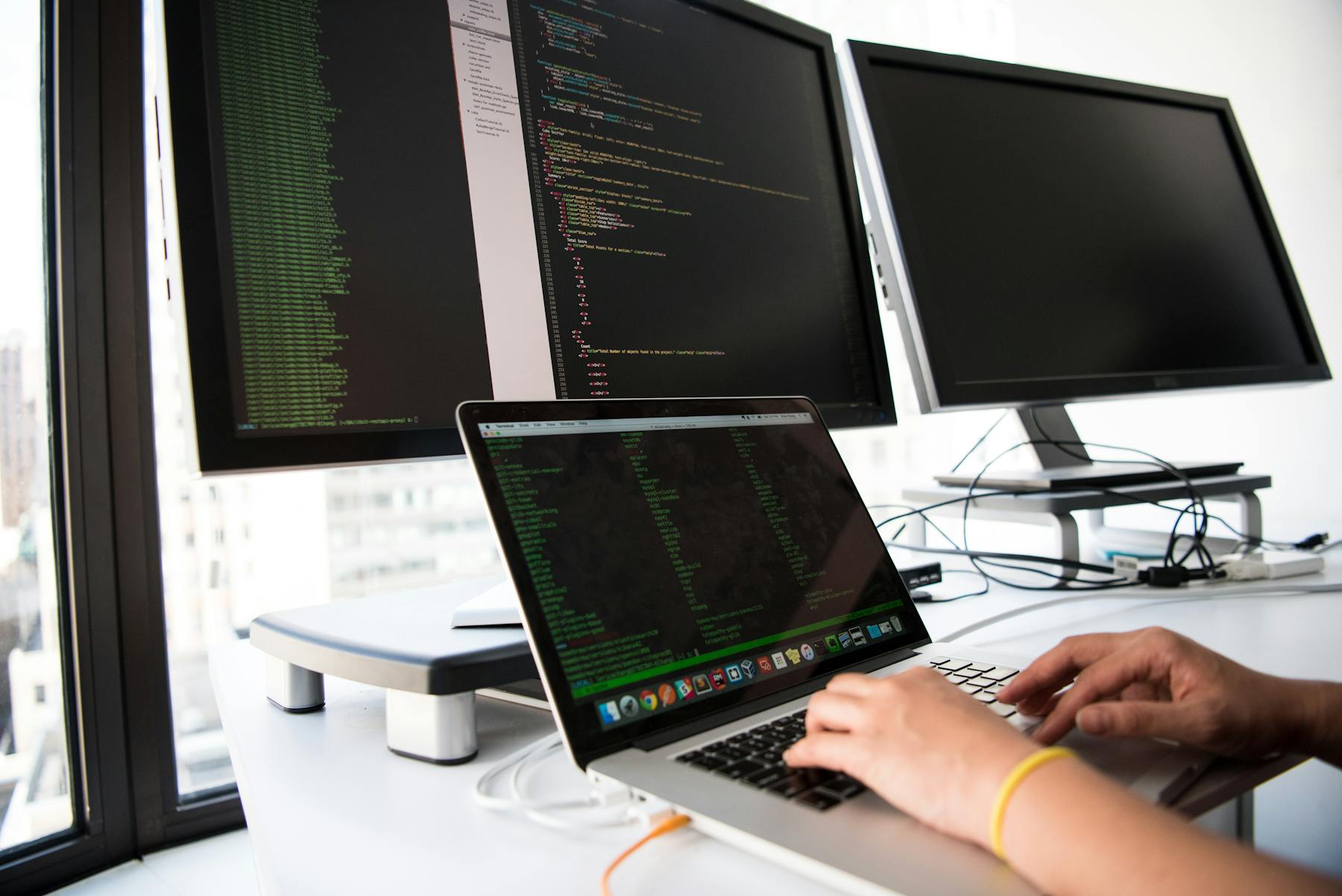 Software developer working with code across a laptop and monitors in a professional workspace