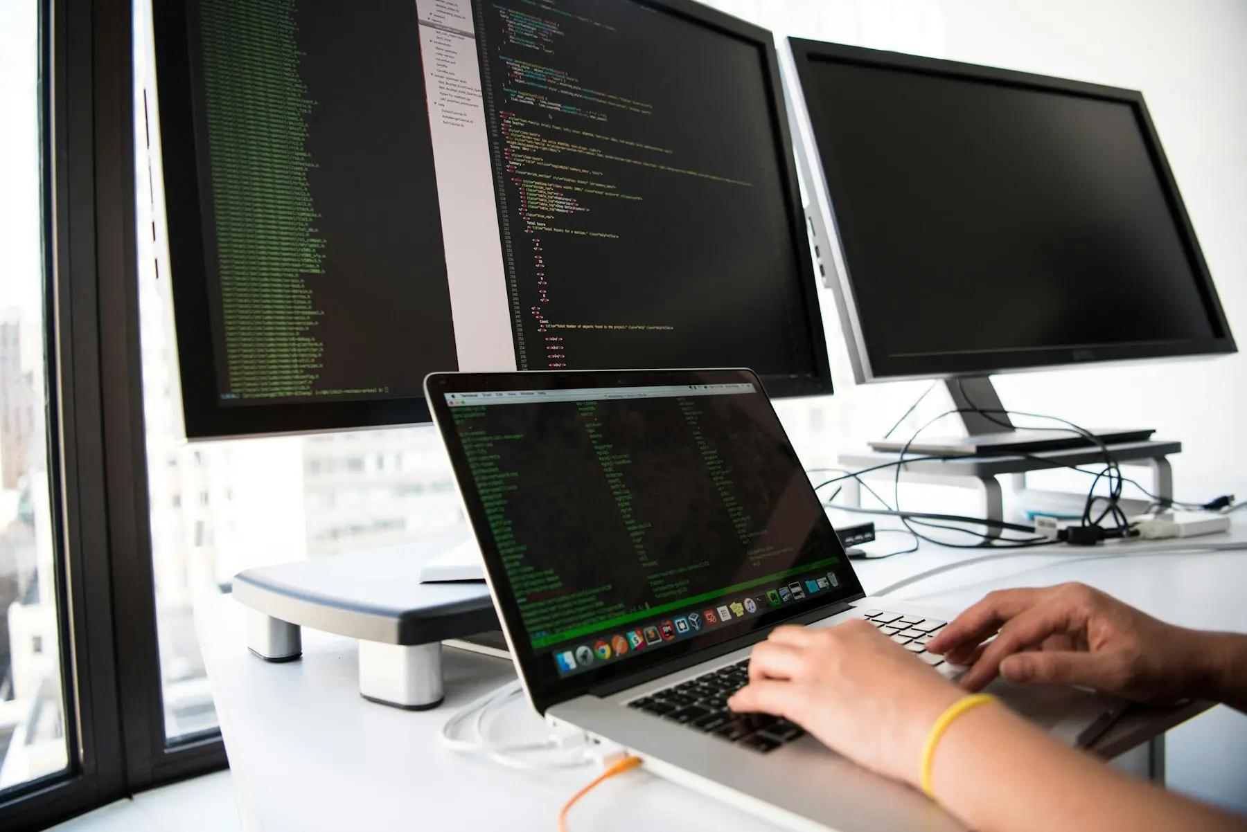 Software developer working with code across a laptop and monitors in a professional workspace